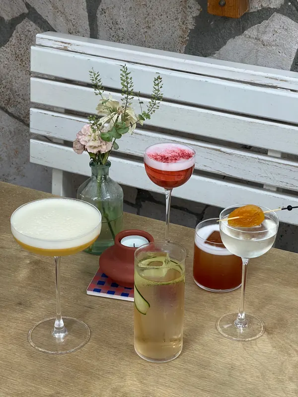 Five cocktails and a vase of flowers on a table by a white bench